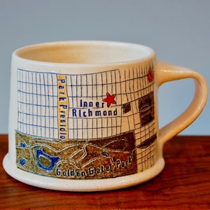 May include: A white ceramic mug with a map of San Francisco printed on it. The map includes the areas of Inner Richmond, Park Presidio, and Golden Gate Park. A red star marks a location on the map.