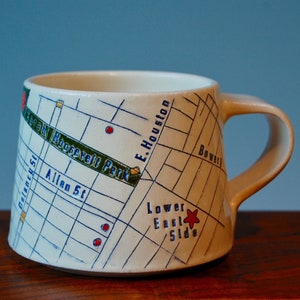 May include: A white ceramic mug with a map of the Lower East Side of Manhattan, New York City. The map includes the street names "E. Houston", "Bowery", "Allen St", and "FDR Drive". The mug also has the words "Lower East Side" and a red star.