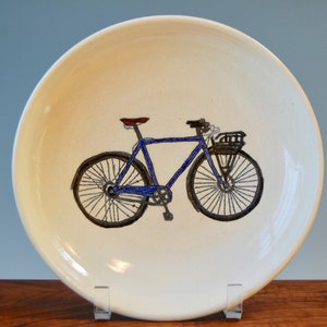 May include: A white ceramic plate with a hand-painted blue bicycle with a brown seat and a black basket. The bicycle has black wheels with white spokes.