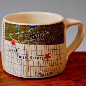 Könnte beinhalten: Eine weiße Keramiktasse mit einem Kartenmotiv. Die Karte zeigt die Stadtteile Judah, Outer Sunset und Noriega in San Francisco. Die Tasse hat einen braunen Henkel und ein grün-blaues Kartenmotiv. Der Text "Polo Golden Gate Park" ist auf der Tasse sichtbar.