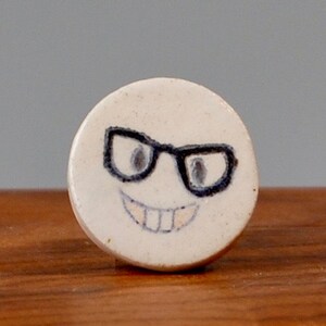 May include: A white ceramic round coaster with a black and white illustration of a smiling face wearing glasses.