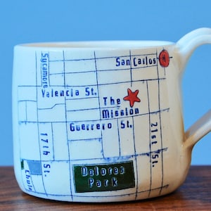 Puede incluir: Una taza de cerámica blanca con una ilustración de mapa en tinta azul del distrito The Mission en San Francisco. El diseño incluye nombres de calles como Valencia St, Guerrero St, 17th St, 21st St y Sycamore, una estrella roja, un círculo rojo y Dolores Park.