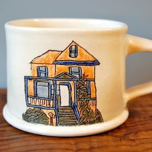 May include: White ceramic mug featuring a hand-painted illustration of a yellow house with a blue roof and a porch. The house has a blue door and windows.