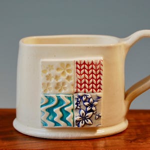 May include: White ceramic mug with a square handle. The mug features a decorative design of four squares with different patterns in blue, red, white, and yellow.