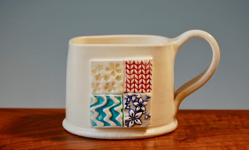 May include: White ceramic mug with a square handle. The mug features a decorative design of four squares with different patterns in blue, red, white, and yellow.