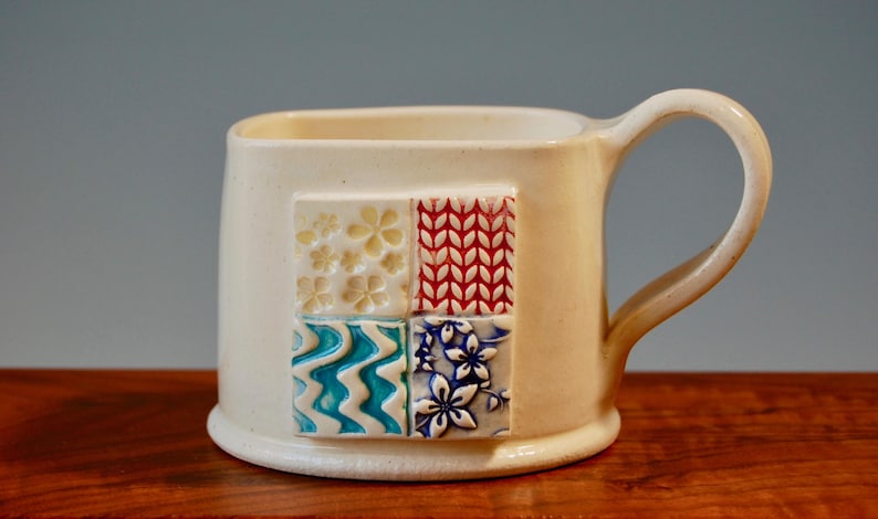 May include: White ceramic mug with a square design featuring four different patterns in blue, red, white, and yellow.