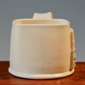 May include: A white ceramic mug with a square shape and a handle. The mug has a subtle green and white design on the side.