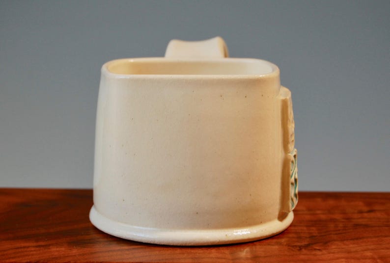 May include: A white ceramic mug with a square shape and a handle. The mug has a subtle green and white design on the side.