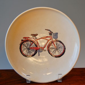 May include: A white ceramic plate with a red bicycle painted on it. The bicycle has a basket on the front and black tires.
