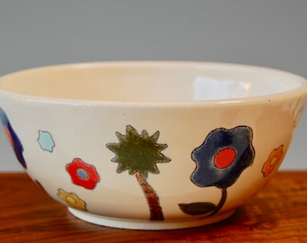 May include: A white ceramic bowl with a floral design. The bowl features blue, red, yellow, and green flowers and a green tree.