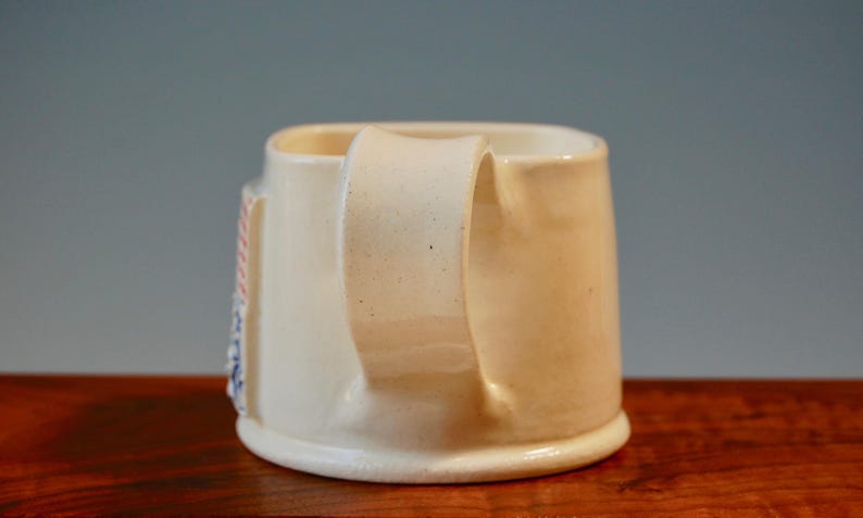 May include: A white ceramic mug with a handle. The mug has a design on the side with red, white and blue stripes.
