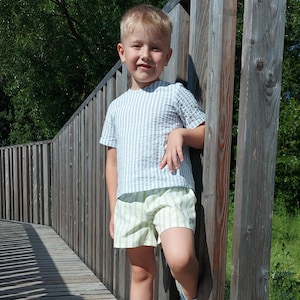 Puede incluir: Un niño pequeño con una camisa a rayas azul claro y blancas y pantalones cortos a rayas verdes y blancas está de pie en una pasarela de madera. Se apoya en una valla de madera y sonríe a la cámara.
