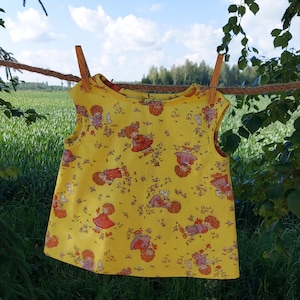 Può includere: Un vestito giallo senza maniche con un motivo di piccole ragazze e fiori. Il vestito è appeso a uno stendino.