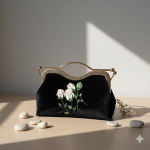 May include: A black velvet handbag featuring an embroidered design of three white rosebuds with green stems and leaves. The bag has a gold-toned metal frame with a curved handle and a textured, decorative edge along the top closure mechanism.