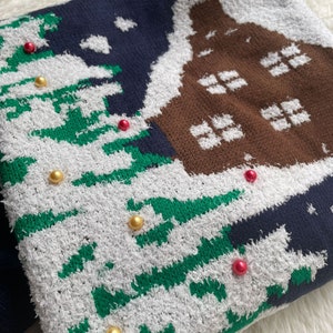 May include: A blue, green, white, and brown knitted sweater with a house and Christmas trees design. The sweater has gold and red beads sewn on it.