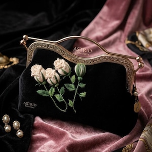May include: Black velvet clutch with a gold-toned frame and handle. The clutch features embroidered cream-colored roses and green leaves. The brand name "ASTRID Embroidery" is visible on the clutch. Pearl earrings are in the lower left corner.
