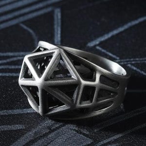 May include: A silver-colored ring with a geometric design. The ring features a complex, openwork structure with multiple facets and angles. The ring is set against a dark background with a subtle pattern.