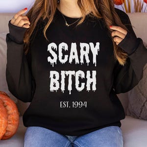 Puede incluir: Sudadera negra con texto blanco que dice "Scary Bitch Est. 1994". El texto está goteando, lo que le da un ambiente espeluznante de Halloween.