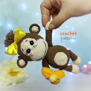 Può includere: Giocattolo di scimmia all'uncinetto con pelliccia marrone e bianca, che tiene una banana gialla. La scimmia ha un fiocco giallo sulla testa e occhi neri. Il testo "crochet pattern" è scritto in arancione su sfondo blu.