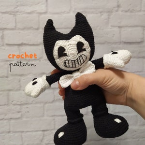 May include: Crocheted black and white Bendy plush toy with a bow tie. The toy is holding out its arms and has a big smile. The text "crochet pattern" is written in orange on the left side of the image.
