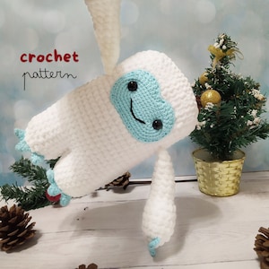 May include: White crocheted snowman amigurumi toy with a blue heart-shaped face and black eyes. The toy is standing on a white wooden surface with a small pine tree in a gold basket behind it. The text "crochet pattern" is written in red and white on the left side of the image.