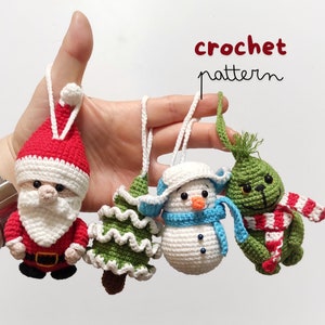 Op de afbeelding: Vier gehaakte kerstversieringen: een rode kerstman, een groene kerstboom, een witte sneeuwpop en een groene Grinch. De versieringen hangen aan een hand.