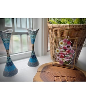 May include: Two decorative blue and grey vases with a floral design stand near a window. A framed Hamsa hand with flowers and text is on a wooden slab. A woven basket with greenery is in the background.