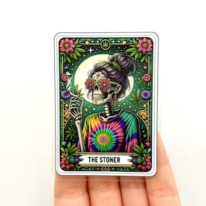May include: A tarot card with a green, pink and purple tie-dye design. The card features a skeleton with a flower crown and sunglasses smoking a joint. The card is titled "THE STONER".