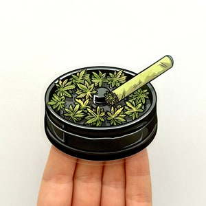 Peut inclure: Autocollant représentant un grinder noir rempli de feuilles de cannabis vertes et un joint allumé. L'autocollant a une bordure blanche et est posé sur un fond blanc.