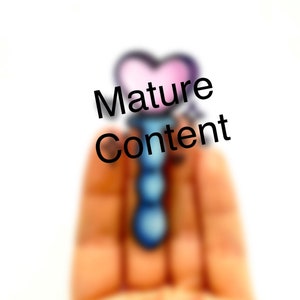 May include: A blurry image of a hand holding a blue and pink heart-shaped charm with the text "Mature Content" written in black.