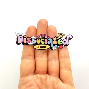 Op de afbeelding: Een transparante sticker met het woord "dissociated" in kleurrijke bubbelletters. De sticker heeft een regenboog, een wolk, een hart, een bloem en een halve maan met de tekst "BRB" erin.
