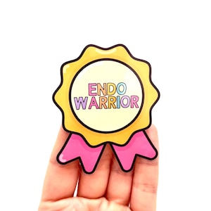 May include: A sticker featuring the text "ENDO WARRIOR" in a rainbow gradient, set within a yellow award ribbon design with a pink bow. The sticker is held in a hand against a white backdrop.