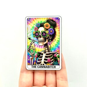May include: A sticker featuring a skeleton with a tie-dye background, wearing sunglasses and a flower crown, smoking a joint. The text "THE CANNABITCH" is printed below the skeleton.