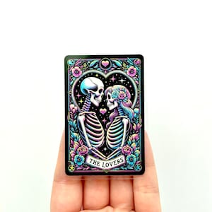 May include: A tarot card with a black background and a gold frame. The card depicts two skeletons embracing, with a heart and stars in the background. The text "THE LOVERS" is printed on a banner below the skeletons.
