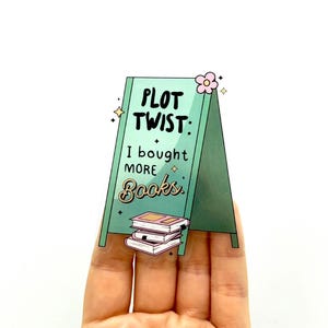 Puede incluir: Un cartel verde y blanco con el texto "Plot Twist: I bought more books." El cartel tiene una flor rosa y brillos. Una pila de libros rosas se encuentra en la parte inferior del cartel.