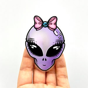 Puede incluir: Una pegatina de cabeza de extraterrestre morada con un lazo rosa y un amuleto de planeta azul. La pegatina tiene un acabado brillante y es perfecta para agregar un toque de fantasía a tu computadora portátil, botella de agua o planificador.