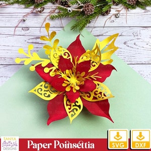 May include: A red and gold paper poinsettia flower with intricate cut-out details. The flower is layered on a light green surface, with text that reads "Paper Poinsettia" and the Tanya Batrak Designs logo. Includes SVG and DXF download icons.
