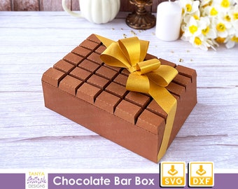 May include: A brown cardboard box shaped like a chocolate bar with a gold ribbon bow. The box is on a white wooden surface. The text "Chocolate Bar Box" is visible below the box. The text "SVG" and "DXF" is visible below the box.