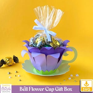 May include: A purple and green paper craft flower cup gift box with a clear cellophane bag filled with assorted chocolates. The cup is sitting on a white saucer.