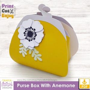 Può includere: Una scatola gialla a forma di borsetta con un fiore di anemone bianco e foglie verdi. L'interno della scatola è bianco e grigio. Il testo sull'immagine recita "Print Cut Enjoy" e "Purse Box With Anemone".