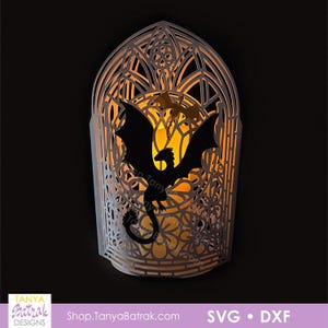 May include: A white laser-cut paper lantern featuring an arched gothic window frame with intricate floral patterns and two dragon silhouettes. The interior is illuminated with a warm orange glow. The text Tanya Batrak Designs Shop.TanyaBatrak.com SVG DXF is visible.