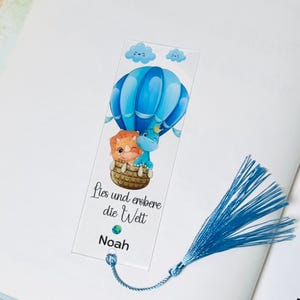 Könnte beinhalten: Ein blauer und weißer Heißluftballon mit zwei Dinosauriern darin. Der Ballon fliegt mit Wolken am Himmel. Das Lesezeichen hat den Text "Lies und erobere die Welt" und "Noah" darauf. Es hat einen blauen Quasten.