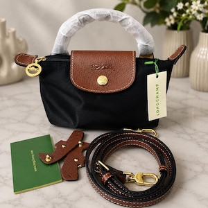 May include: A black nylon handbag with a brown leather flap featuring a gold-tone snap button and an embossed horse logo. The bag has a white paper tag labeled LONGCHAMP, a detachable brown leather shoulder strap, and a green informational card.