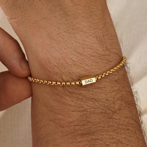 Puede incluir: Una pulsera de cadena tipo caja en tono dorado que presenta una pequeña cuenta metálica rectangular y horizontal centrada en la cadena. La cuenta está grabada con la palabra DAD en letras mayúsculas. La pulsera consta de eslabones redondeados.