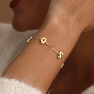Op de afbeelding: Een delicate gouden kettingarmband met twee kleine, gepolijste gouden letterbedels, de letters O en S, gelijkmatig verdeeld over de dunne metalen schakelketting. De armband heeft een heldere, reflecterende afwerking en een eenvoudig ontwerp.