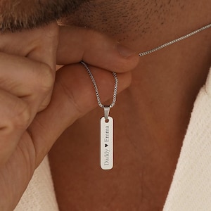 Könnte beinhalten: Eine silberfarbene Halskette mit einem vertikalen Stab-Anhänger an einer feinen Kette. Der rechteckige Metallanhänger ist mit dem Text "Daddy ♥ Emma" in einer klaren, serifenlosen Schrift graviert. Der Anhänger hängt an einer zarten Kette.