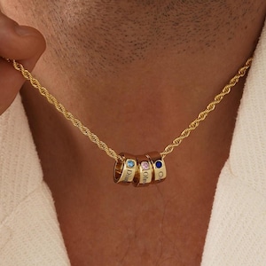 May include: A gold-toned rope chain necklace featuring three small, cylindrical charms. Each charm is engraved with a name and set with a single, small round gemstone in blue, pink, and dark blue, suspended from the centre of the chain.