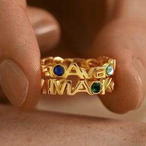 May include: Two gold-colored rings with the names "Ava" and "Mack" spelled out in capital letters. Each ring has a different colored gemstone: blue, green, and teal. The rings are held between two fingers.