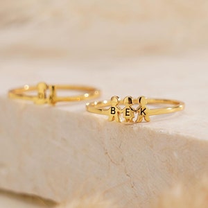 May include: Two gold rings are shown, one in the background and the other in the foreground. The foreground ring has three small figures with the letters "B", "E", and "K" incorporated into the design. The rings are set against a neutral, textured background.