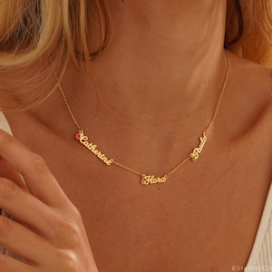 May include: Gold necklace with three names, "Catherine", "Flora", and "Paula", spelled out in cursive with a small red gemstone on the "Catherine" charm, a small green gemstone on the "Paula" charm, and a small purple gemstone on the "Flora" charm.